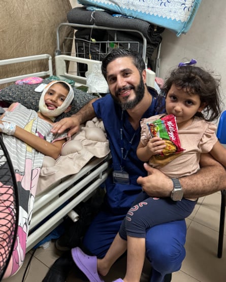 A girl lies in a hospital bed with a bandage around her badly damaged face. A doctor sits next to her with a younger girl on his lap