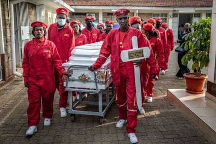 Young men and women, all dressed in red jumpsuits and red berets, walk pushing a white casket on a trolley