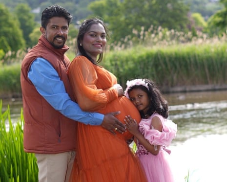 A pregnant woman in an orange dress stands with a man and young girl beside a pond