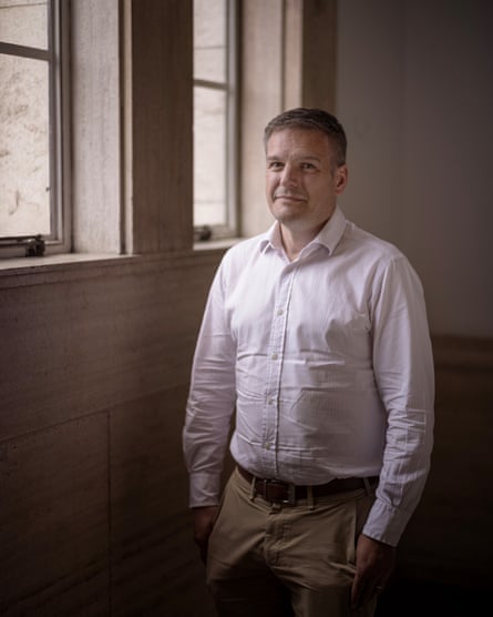 Brian bell stands with his hands in his pockerts for a photograph, lit by light from a window