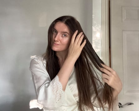 The writer is in a white dressing gown in a white room, running product through her long brown hair. She is facing the camera.