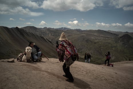 A man in traditional dress takes a photo of a tourist and an alpaca against a mountain backdrop