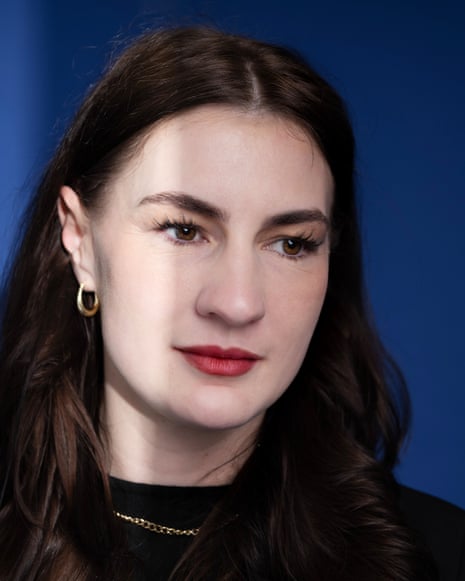 A close-up of Lucia Osborne-Crowley, her dark hair in a middle parting, tucked around one ear to show a gold hoop. She smiles very gently, looking to her left, away from the camera