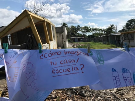 A sign hangs on a line, held on with pegs, with the words “How do you imagine your home/school?” in Spanish. In the background are damaged buildings