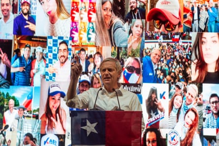 A man speaks at a lectern draped in a Chilean flag against a colourful backdrop of images of members of the public