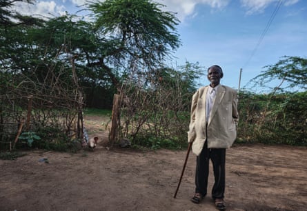 An old African man leaning on a stick stands in a traditional compound with a large tree in the background