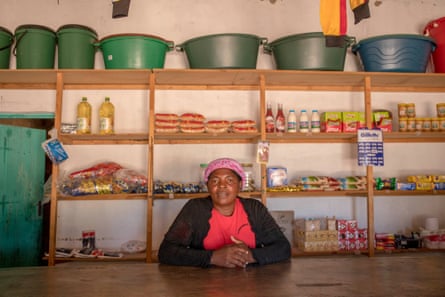 An older woman sits behind a shop counter with a few shelves sparsely filled with goods behind her