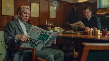 Two men in a grey suits with grey hair sitting together in a wood panelled office looking down at their newspapers