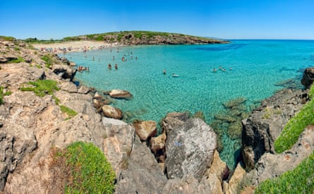 A turquoise bay with some swimmers in it with a small sandy beach and rocky surroundings