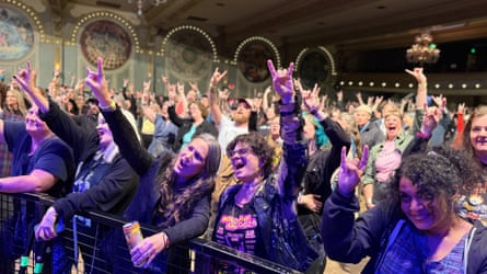 A crowd of people cheer and hold up their pointer and pinky fingers