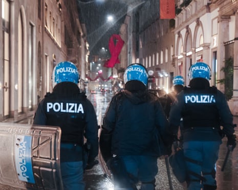 Police officers in riot helmets and shields walk down a rainy Milan street at night