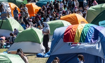 Dozens of tents and young protesters wearing keffiyeh, with a rainbow Pride flag in the foreground.
