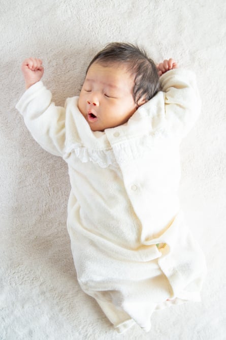 A baby lying on a white rug wearing a white sleep suit and yawning