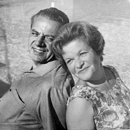 Black and white photo of a man and woman sitting back to back and smiling. He is wearing a collared shirt and she is wearing a sleeveless floral dress
