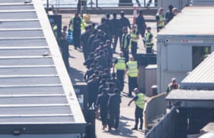 Workers in hi-vis vests shepherd a queue of people all dressed in black