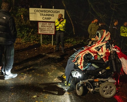 A man in a wheelchair wearing a union flag rain poncho among people standing in the dark by a sign for Crowborough training camp