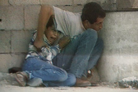 A man protects his crying preteen boy, as they both hide behind a barrel in the street