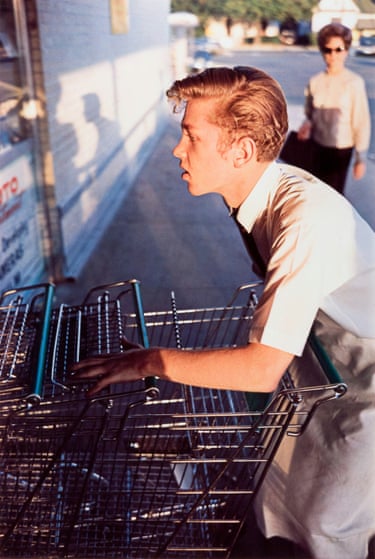 A young boy with a quiff, wearing an aron, stacking supermarket trollies. A lady wearing sunglasses is in the background