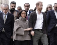 The Duchess of Sussex in a funnel-neck jacket holding hands with her husband and surrounded by men in Jordan