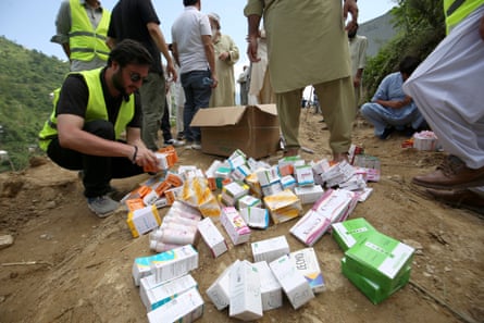 A man in a hi-vis vest crouches next to a collection of medicine boxes lying on the muddy ground. Several other people can be seen standing behind him.