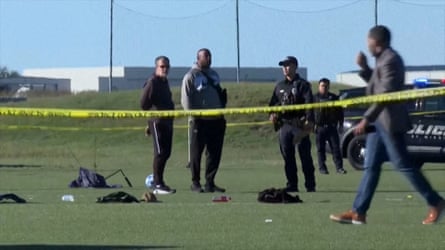 Police and other people stand outdoors on soccer field behind yellow police tape