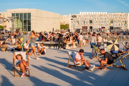 A sunny terrace with many suntanned young people sitting on deckchairs and the colourful exterior of a blocky art gallery in the background