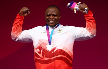 Tchatchet, wearing a Team England tracksuit, smiles and raises his arms while wearing his medal on the podium