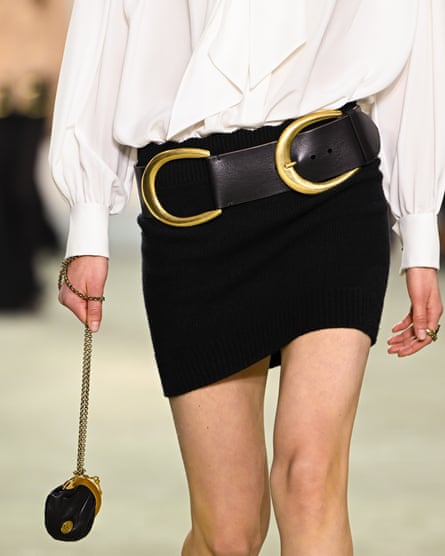 Midshot below neck and above knees shows model carryng small bag on chain in white blouse with black skirt and oversized belt with double gold buckle