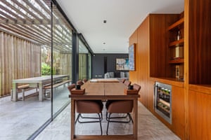 A six-seater table with an integrated wooden storage unit and wine cooler on onside and floor to ceiling glazing on the other. On the other side of the glass it a large blocky table with wood-topped benches