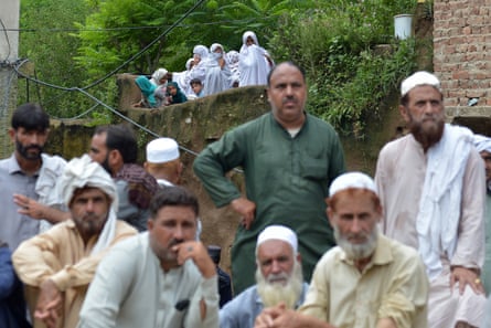A group of men, out of focus in the foreground, all look in the same direction. A group of women and children are standing above and behind them, in focus. Plants and trees are around and behind them