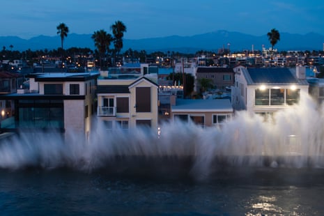 Large waves crash over a seawall against Long Beach houses at dusk, palm trees behind