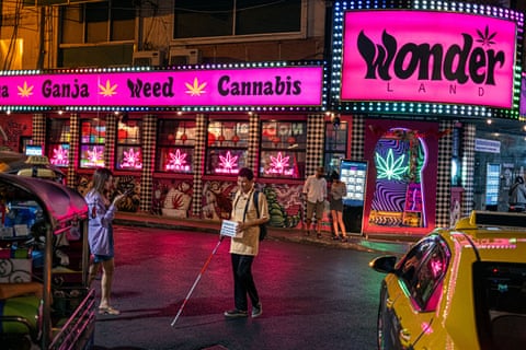 People look at their phones outside the 'Wonder Land' cannabis shop as a blind man using a white and red cane walks past with a donation box hanging around his neck