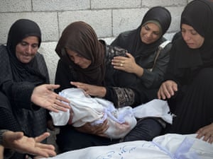 A weeping woman in a chador cradles the body of a small child wrapped in a white sheet as three others around her comfort her