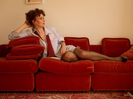 Portrait of Miranda July lying on a red sofa wearing an oversized shirt and tie that’s been left undone, with her front to the camera