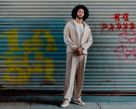 Colin Kaepernick wears smart-casual clothing in neutral colours and stands in front of a corrugated metal sheet with graffiti on it