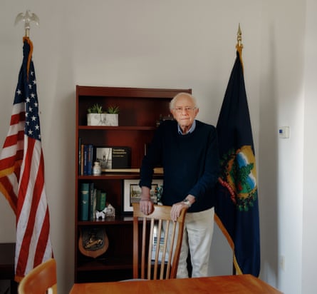 Bernie Sanders standing in his office in front of a table, holding on to the back of a chair, with a bookcase behind him flanked by the US and state flags
