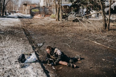 An injured, bloodied man lies on a war-torn street beside bags, surrounded by fallen branches and debris
