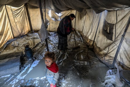 Woman tries to sweep out filthy grey water and mud from inside tent with small child in foreground