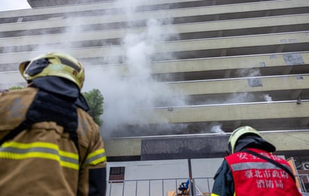 Smoke billows in front of a multi-storey building as rescue workers stand below with their backs to the camera