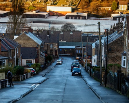 View down a hilly residential street of mostly terrace brick houses, showing steelworks buildings with corrugated metal roofs in the background at the bottom; the wet road is glistening
