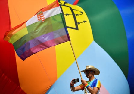 A man wearing a rainbow-striped top and a cowboy hat holds an LGBTQ+ flag in front of a huge Pride flag.