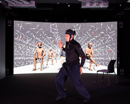 A performer wearing a motion-capture headset and dark clothing moves across a studio stage in front of a large curved screen displaying digital human figures seated against an abstract black-and-white background.