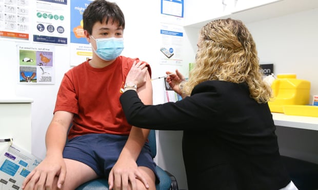 A young boy wearing a face mask about to receive a dose of a Covid vaccine. A pharmacist sits next to him holding a needle.