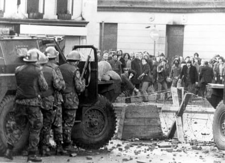 A black and white photo of armed soldiers shielding behind a military vehicle while a group of protesters stand back in the street.