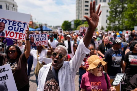Shot of crowds marching and holding banners, with man in foreground chanting with palm raised.