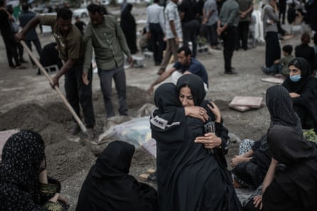 Women dressed in black hug each other as a gravedigger in the background digs a hole in the ground