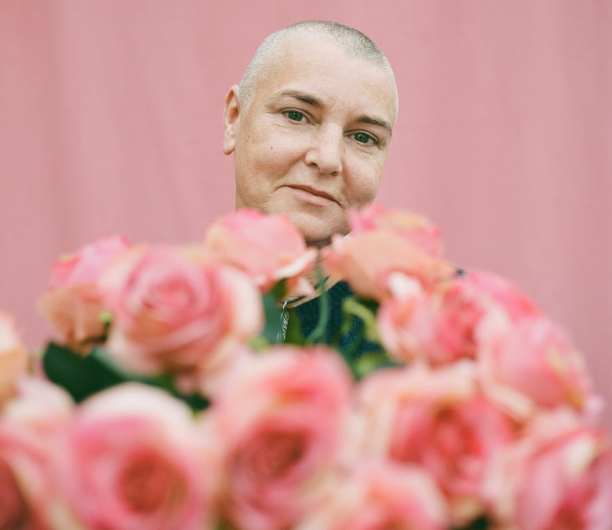 Head shot of Sinéad O'Connor against pink background, with a bunch of pink roses in front of her
