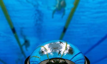 An underwater view shows swimmers reflected in the lens of an underwater robotic camera