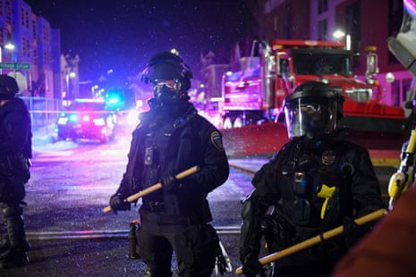 Police officers in full riot gear hold their batons in both hands on a wintery street that is lit up by police lights.