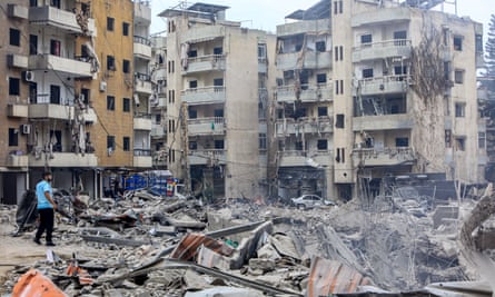 A man stands at the edge of a rubble-strewn area surrounded by blast-damaged buildings